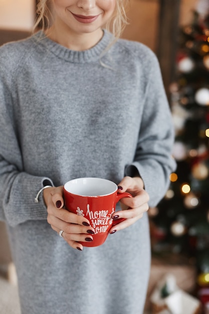 Uma bela jovem com um suéter aconchegante mantém um copo de bebida quente e posando perto da árvore de natal no interior decorado para o ano novo