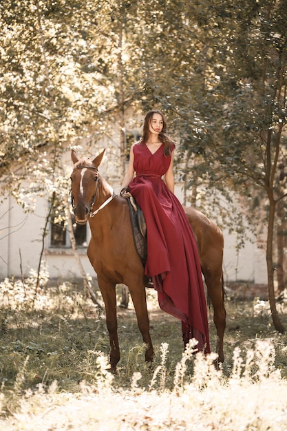 Uma bela jovem com um vestido de seda montada em um cavalo. Passeio a cavalo em um dia ensolarado na floresta.