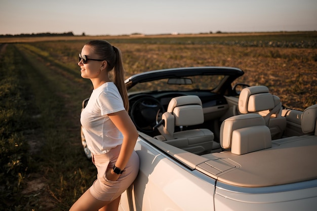 Uma bela jovem de aparência europeia no rosto de óculos de sol, ela fica perto de seu conversível branco. mulher de negócios bem sucedida e seu carro branco.