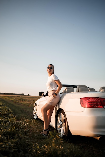 Uma bela jovem de aparência europeia no rosto de óculos de sol, ela fica perto de seu conversível branco. Mulher de negócios bem sucedida e seu carro branco.