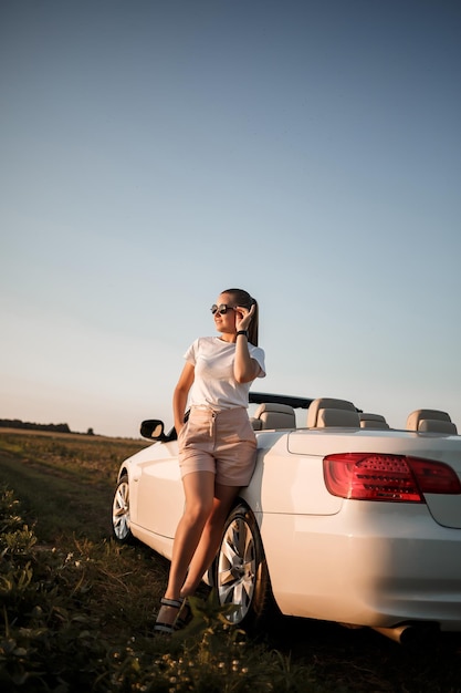 Uma bela jovem de aparência europeia no rosto de óculos de sol, ela fica perto de seu conversível branco. Mulher de negócios bem sucedida e seu carro branco.