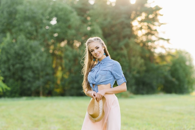 Uma bela jovem de camisa jeans e saia rosa tem um chapéu nas mãos, ela sorri e caminha pela natureza no verão