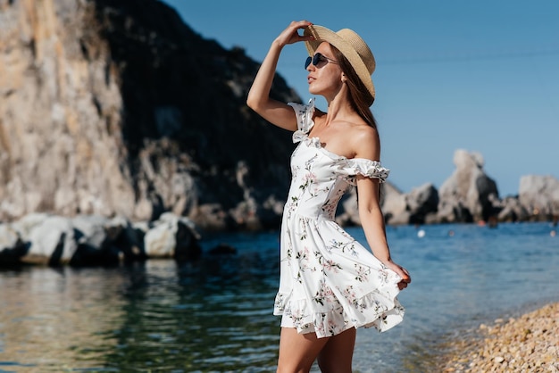 Uma bela jovem de óculos de chapéu e um vestido leve está andando ao longo da costa do oceano no contexto de enormes rochas em um dia ensolarado Turismo e viagens turísticas