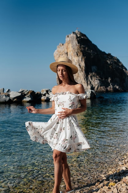 Uma bela jovem de óculos de chapéu e um vestido leve está andando ao longo da costa do oceano no contexto de enormes rochas em um dia ensolarado Turismo e viagens turísticas