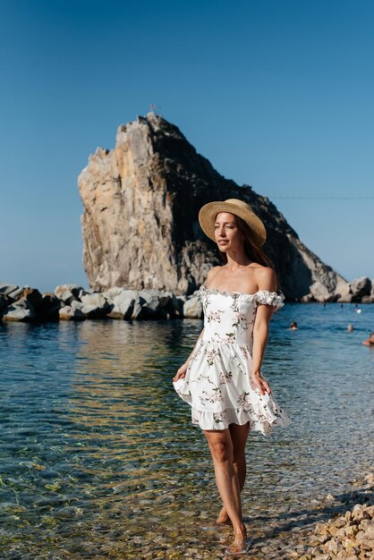 Uma bela jovem de óculos de chapéu e um vestido leve está caminhando ao longo da costa do oceano no contexto de enormes rochas em um dia ensolarado Turismo e viagens turísticas