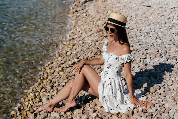 Uma bela jovem de óculos de chapéu e um vestido leve está sentada de costas para o oceano no contexto de enormes rochas em um dia ensolarado Turismo e viagens turísticas