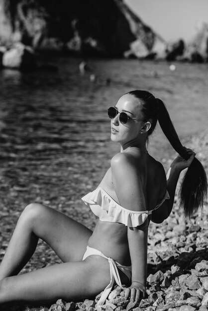 Uma bela jovem de óculos e biquíni está sentada na costa do oceano no contexto de pedras enormes em um dia ensolarado. Turismo e viagens turísticas. Preto e branco.