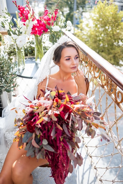 Uma bela jovem em lingerie requintada fica na varanda de um prédio de vários andares e tem um grande buquê de lindas flores nas mãos manhã da noiva no dia do casamento