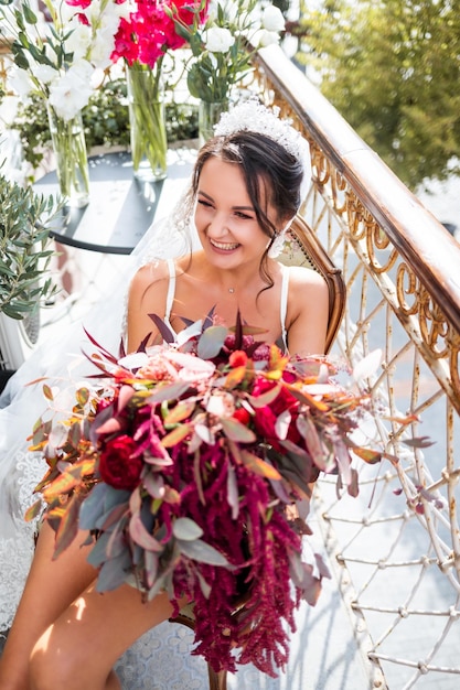 Uma bela jovem em lingerie requintada fica na varanda de um prédio de vários andares e tem um grande buquê de lindas flores nas mãos Manhã da noiva no dia do casamento