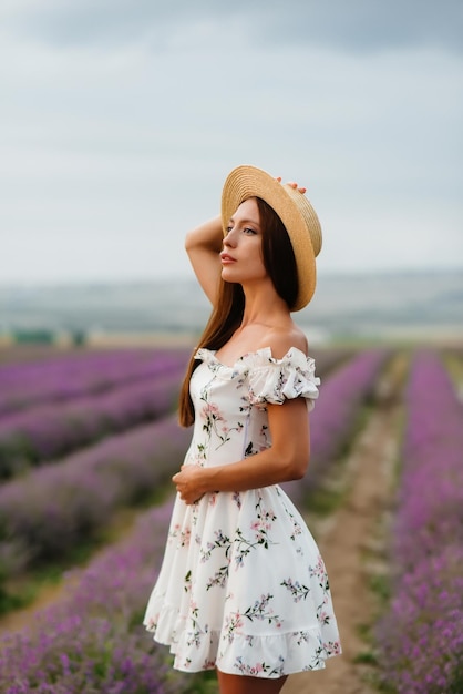 Uma bela jovem em um vestido delicado e chapéu caminha por um belo campo de lavanda e aprecia o aroma das flores. Férias e beleza natural.
