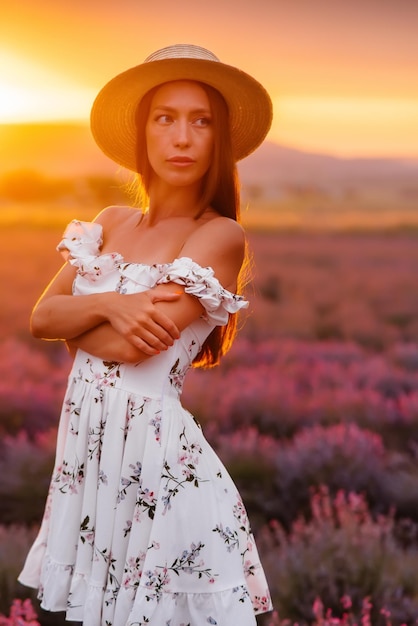 Uma bela jovem em um vestido delicado e chapéu caminha por um belo campo de lavanda e aprecia o aroma das flores. Férias e beleza natural.