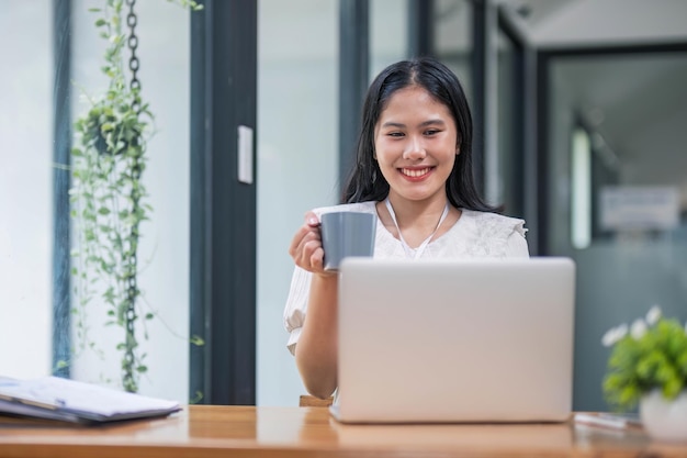 Uma bela jovem empresária asiática a beber café a trabalhar num portátil no escritório.