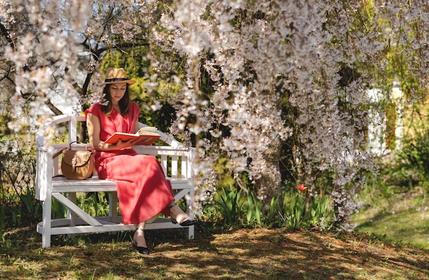 Uma bela jovem está sentada em um banco elegante em um jardim de primavera sob uma flor de cerejeira e lendo um livro