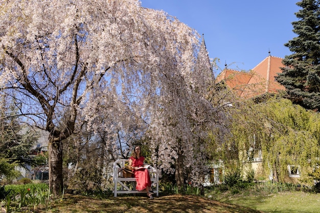 Uma bela jovem está sentada em um banco elegante em um jardim de primavera sob uma flor de cerejeira e lendo um livro