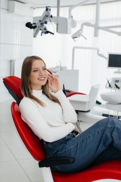 Uma bela jovem está sentada em uma cadeira odontológica vermelha na odontologia branca moderna e sorrindo Tratamento de próteses dentárias e clareamento dos dentes