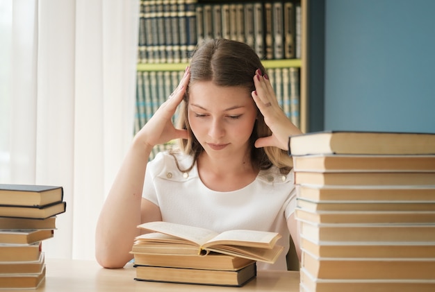 Uma bela jovem estudante está sentada na biblioteca entre enormes pilhas de livros