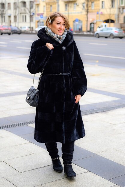 Uma bela jovem feliz com um elegante casaco de pele de inverno caminhando por uma rua da cidade