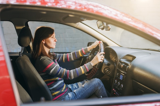 Uma bela jovem feliz e sorridente mulher europeia de cabelos castanhos com pele limpa e saudável, vestida com uma camiseta listrada, senta-se em seu carro vermelho com interior preto. viajar e dirigir o conceito.