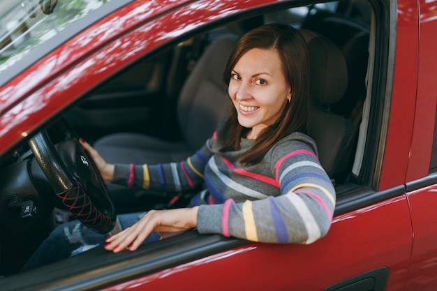 Uma bela jovem feliz e sorridente mulher europeia de cabelos castanhos com pele limpa e saudável, vestida com uma camiseta listrada, senta-se em seu carro vermelho com interior preto. Viajar e dirigir o conceito.