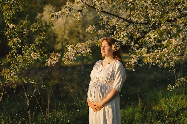 Uma bela jovem grávida em um vestido branco caminha no jardim da primavera