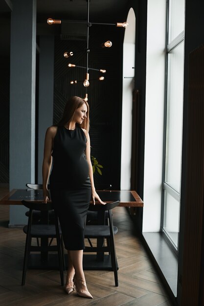 Uma bela jovem grávida em um vestido preto está posando no interior escuro de um restaurante.