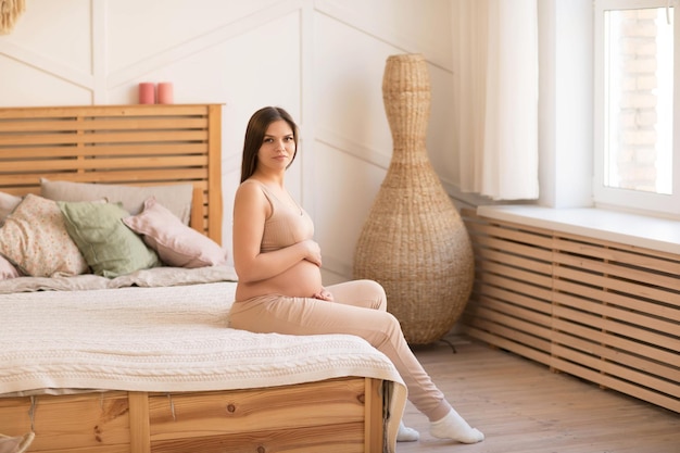 uma bela jovem grávida senta-se em uma cama perto da janela e olha para a câmera
