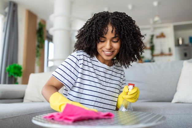 Uma bela jovem mulher faz a limpeza da casa, uma menina esfrega a poeira, limpa a mesa e pulveriza desinfetantes, usa soluções de limpeza ou usa álcool para matar germes.