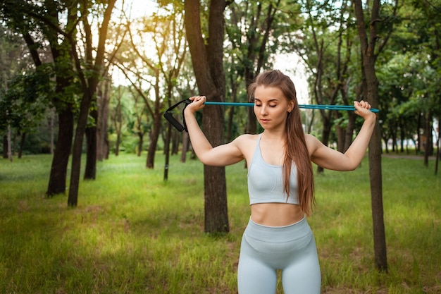 Uma bela jovem realiza um exercício ao ar livre. Grama, parque, treino de esportes. Ginástica. Um expansor. Equipamento desportivo, pôr do sol. Um estilo de vida saudável, corpo musculoso treinado. Floresta na cidade