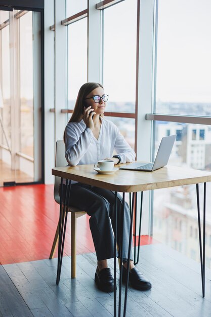 Uma bela jovem ri enquanto fala ao telefone enquanto está sentado em um café fazendo um pedido e anotando em um laptop Garota positiva ligando pelo celular enquanto está sentado com café