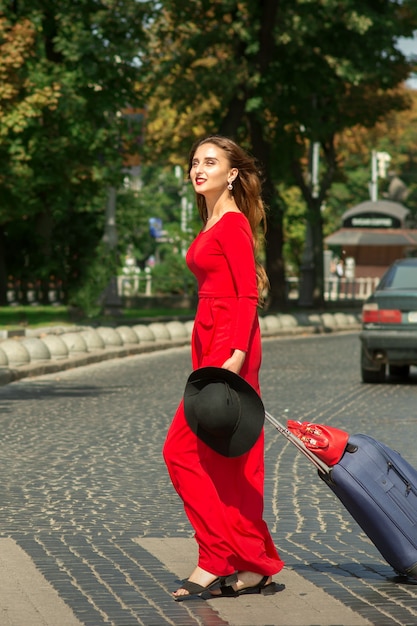 Uma bela jovem turista caucasiana com uma mala em um vestido longo vermelho atravessa a rua em uma faixa de pedestres na rua da cidade ao ar livre