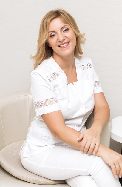 Uma bela médica esteticista de uniforme branco sorri, olha para a câmera e posa enquanto está sentada em uma cadeira em seu escritório. Foto vertical