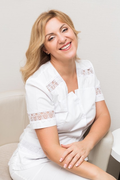 Uma bela médica esteticista de uniforme branco sorri, olha para a câmera e posa enquanto está sentada em uma cadeira em seu escritório. Foto vertical