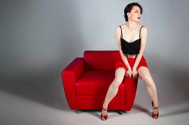 uma bela morena com sapatos vermelhos se senta em uma poltrona vermelha sobre um fundo cinza.