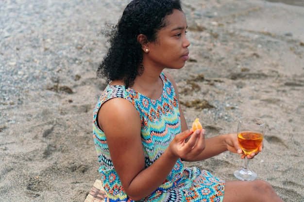 Uma bela mulher africana bebe vinho na praia, olhando para o horizonte.