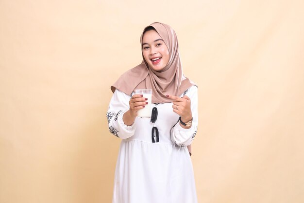 Uma bela mulher muçulmana asiática vestindo um hijab com uma expressão feliz carrega e aponta para um copo