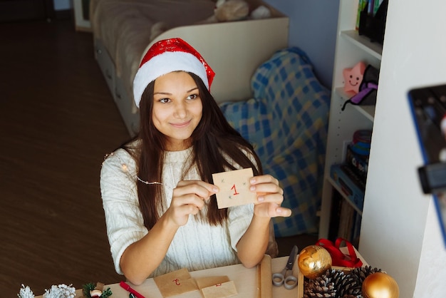 uma blogueira sorridente com um chapéu de Natal cria um calendário do advento e tira fotos em seu telefone