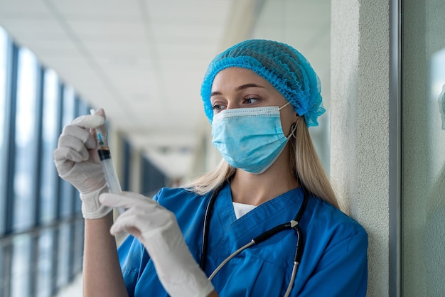 Uma boa enfermeira de uniforme com um estetoscópio nas luvas e uma máscara tem uma seringa nas mãos