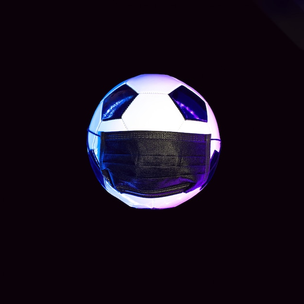 Uma bola de futebol com uma máscara médica preta do vírus. À luz do neon em uma parede escura