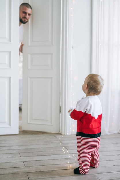 Uma bonitinha menina vestida de Papai Noel de pé na grande sala brilhante olhando para o pai.