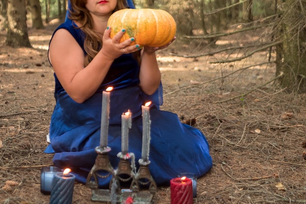 Uma bruxa em um manto azul diz a sorte sobre velas na floresta com uma abóbora nas mãos