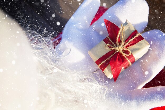 Uma caixa com um presente de Natal nas mãos do Papai Noel em luvas brancas. Terno vermelho, barba, luzes de guirlanda em um borrão. Ano novo, preparação, expectativa de um milagre, um sonho realizado. Fechar-se