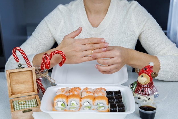 Uma caixa de Filadélfia rola sobre a mesa da cozinha Sushi de entrega rápida em um recipiente branco A menina coloca uma aliança no dedo Decoração festiva Conceito de Natal
