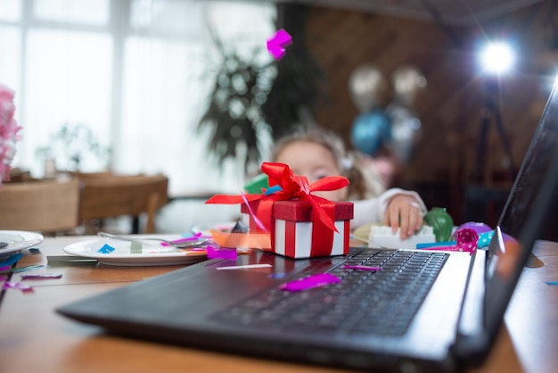 Uma caixa vermelha fica perto do laptop na mesa, muitos confetes coloridos A garota olha para o presente conceito de celebração de aniversário