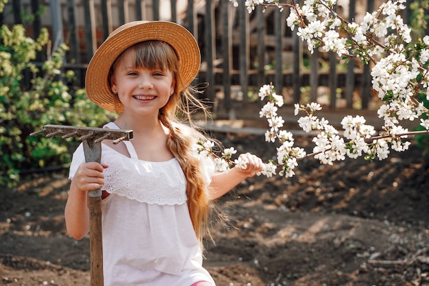 Uma camponesa de chapéu no jardim está envolvida na jardinagem de um ancinho para colher as folhas em suas mãos floração de damascos na primavera o jardim está em flor