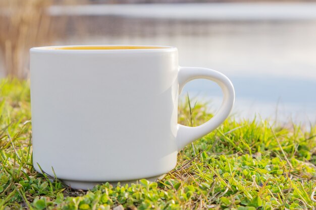 Uma caneca branca fica na grama perto do rio.