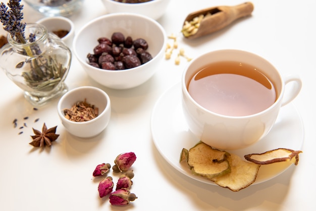 uma caneca branca sobre uma mesa branca com chá de ervas e ingredientes à base de plantas dispostos em cima da mesa. Conceito sobre o tema do tratamento à base de plantas para gripes e resfriados no outono.