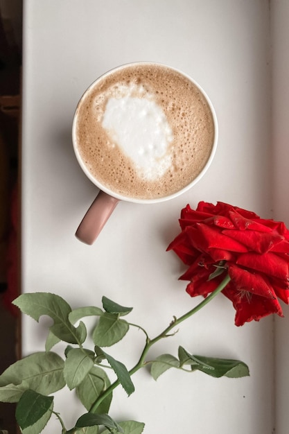 Uma caneca de cappuccino rosa e uma rosa vermelha de jardim estão no parapeito da janela Café da manhã festivo aconchegante em casa vista superior