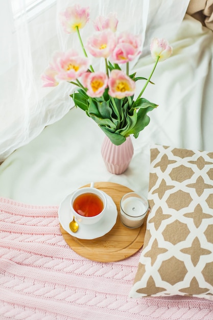 Uma caneca de chá quente, uma manta de tricô rosa, um buquê de tulipas em um vaso, uma vela na cama. Café da manhã na cama. Acolhedor.