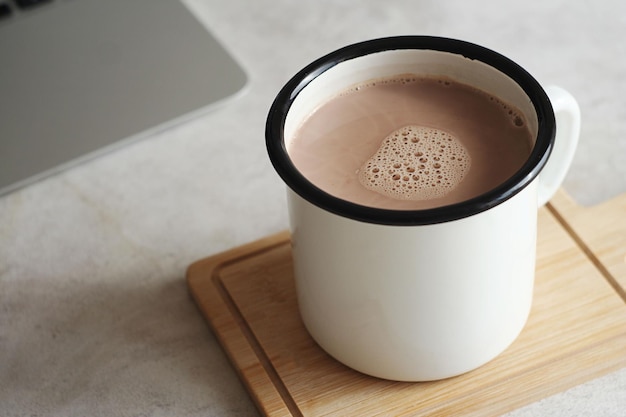 Uma caneca de chocolate quente fica em um suporte de madeira na mesa Atrás está um laptop