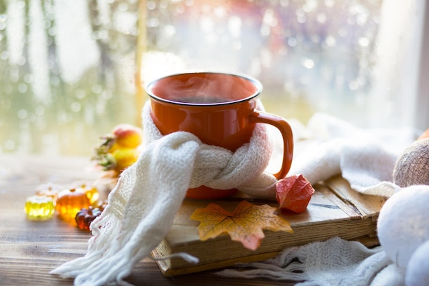 Uma caneca laranja em um cachecol com abóboras de chá quente, maple seco amarelo deixa um livro nas gotas de chuva do peitoril da janela no humor do outono